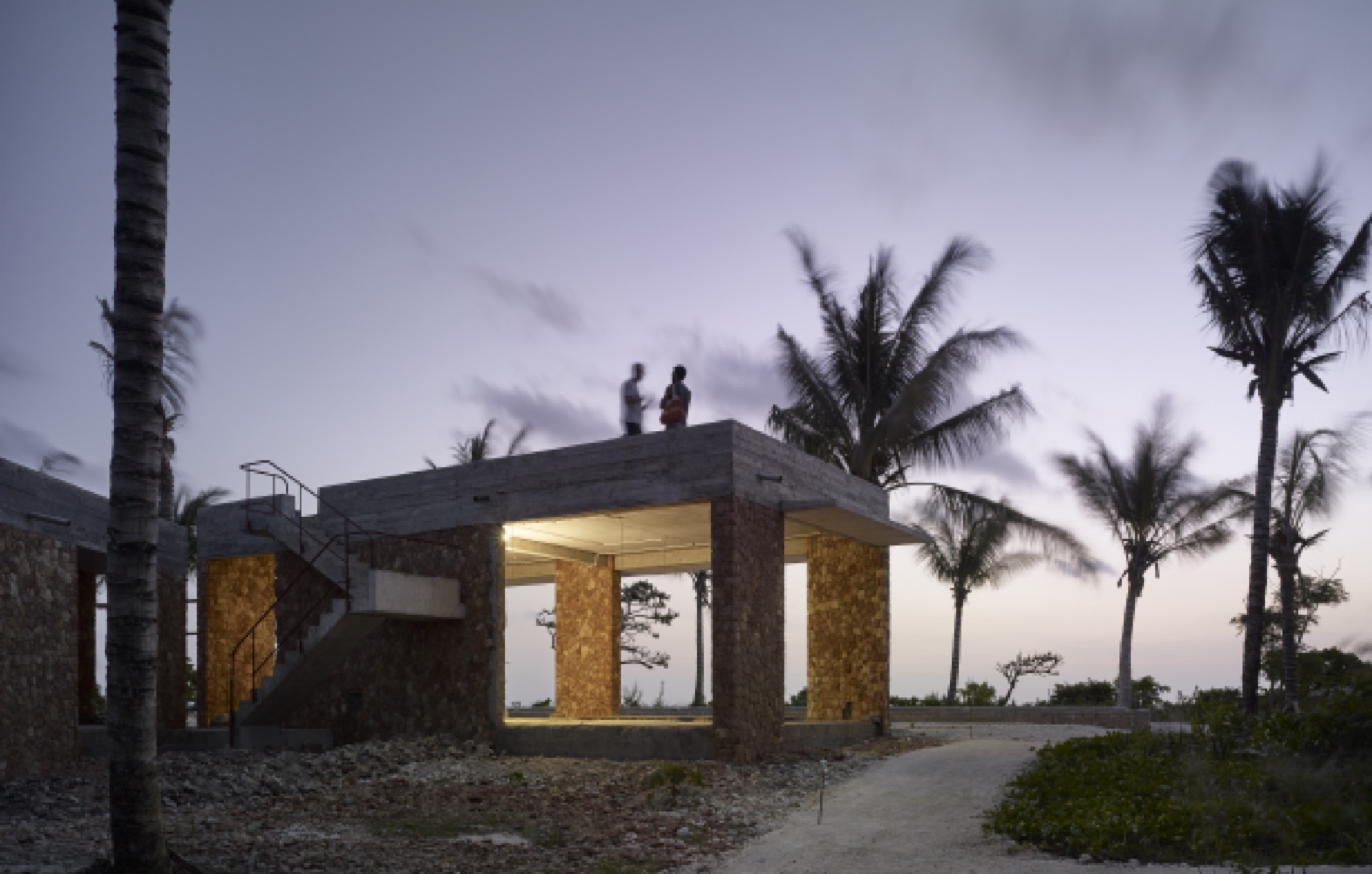 two friends on the rooftop of a coral-stone pavilion at dusk, palms all around